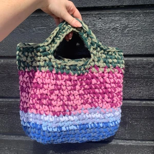 Flerfärgad virkad handväska - Unik handväska med breda ränder i grönt, rosa, lila och blått. Väskan har ett mjukt. Perfekt för dig som vill ha en färgglad accessoar som sticker ut. Stor nog att få plats med de mesta 