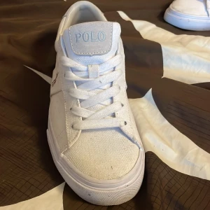 Vita sneakers från Polo Ralph Lauren - Fräscha vita sneakers från Polo Ralph Lauren med klassisk logga på sidan och POLO-broderi på plösen och hälen. Skorna har snörning, rund tå och är tillverkade i canvas med detaljer i mocka. Perfekta för en clean och stilren look.