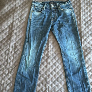 Blå jeans från Jack & Jones - Säljer ett par klassiska blå jeans från Jack & Jones i modellen Clark. Jeansen har fem fickor, snygga slitningar och raka ben. De är tillverkade i bomull och har normal passform med medelhög midja. Perfekta för en avslappnad och stilren look.