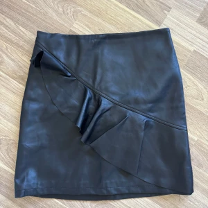 Svart skinnkjol med volang  - Svart kort skinnkjol från Skill med snygg volangdetalj framtill och dragkedja bak. Kjolen har en clean look med en cool asymmetrisk volang som ger extra edge. Perfekt för dig som gillar en stilren men ändå trendig vibe.