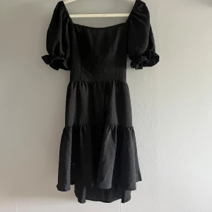 Svart klänning med puffärmar och knyt - Supersöt svart klänning med puffiga kortärmar, volangkjol och snygg knytning i ryggen som ger en öppen rygg. Klänningen har en lätt struktur i tyget och en romantisk vibe med sin feminina siluett. Perfekt för dig som gillar trendiga detaljer och vill sticka ut.