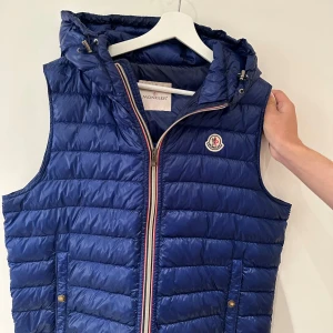 Blå dunväst från Moncler med huva - Snygg blå dunväst från Moncler med quiltad design och avtagbar huva. Västen har dragkedja framtill med röd, vit och blå detalj, två fickor med tryckknappar och Moncler-logga på bröstet. Perfekt för lager-på-lager och streetstyle.