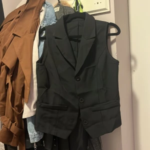 Svart kostymväst med knappar - Stilren svart väst med klassisk kostymkänsla, djup v-ringning och två knappar framtill. Västen har två fickor och breda slag, perfekt för en dressad look. Passar snyggt över skjorta eller t-shirt för en trendig outfit.