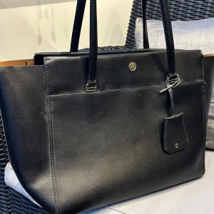 Svart axelväska från Tory Burch - Stilren svart axelväska från Tory Burch i slätt skinn med guldiga metalldetaljer och logga framtill. Väskan har långa handtag, rymligt fack och en snygg tag med logotyp. Perfekt för dig som vill ha en klassisk och tidlös look.