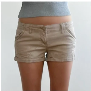 Beige shorts med uppvikta ben - Snygga beige shorts med klassisk femficksdesign och uppvikta ben. De har bälteshällor, knapp och dragkedja framtill samt en avslappnad passform. Perfekta för varma dagar och enkel att matcha med olika toppar.