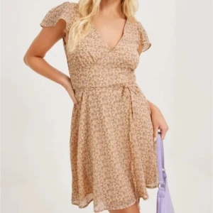 Blommig kortklänning från NLY Trend - Supersöt kortklänning med småblommigt mönster i ljusa färger från NLY Trend. Klänningen har v-ringning, korta volangärmar och knytband i midjan. Lapparna kvar pga aldrig använd OBS har även samma klänning i strl 38