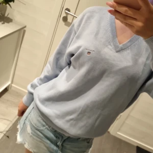 Ljusblå stickad tröja från GANT - Snygg ljusblå stickad tröja från GANT med v-ringad hals och broderad logga på bröstet. Tröjan har långa ärmar och ribbade muddar vid ärmslut och nederkant. Perfekt för dig som gillar klassisk och clean stil.