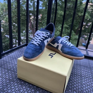 Blå mockasneakers från J.Lindeberg - Snygga blå sneakers från J.Lindeberg i mocka med vita detaljer och vit snörning. Skorna har en klassisk gummisula i beige och en stilren design med rund tå. Perfekta för dig som gillar en clean och sportig look.
