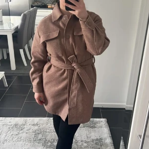 Beige kappa från ONLY - Snygg beige kappa från ONLY med bälte i midjan och stora bröstfickor. Jackan har knappar framtill, klassisk krage och långa ärmar. Perfekt för lager-på-lager och ger en chill men ändå uppklädd vibe. Materialet känns mjukt och har en ullig look.