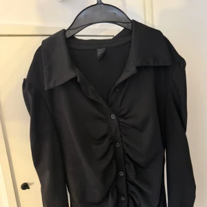 Svart skjorta med rynkade detaljer - Svart långärmad skjorta från Shein med snygga rynkade detaljer längs knappraden framtill. Skjortan har klassisk krage och knappar hela vägen. Perfekt för dig som vill ha en stilren men ändå trendig look. Använd endast få gånger.