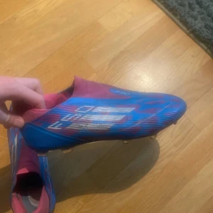 Adidas F50 blå/rosa fotbollsskor - Säljer ett par Adidas F50 fotbollsskor i en riktigt snygg blå och rosa färgkombination med vita detaljer. Skorna har en strömlinjeformad design och är tillverkade i syntetmaterial för bästa känsla på planen. En skadad sluta men lätt och billigt att köpa nya. Perfekta för dig som vill sticka ut på planen! Nypris 1200 mitt pris 499!!