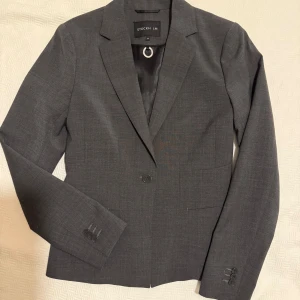 Mörkgrå kavaj från STOCKH LM, stl 34 - Snygg mörkgrå kavaj från STOCKH LM i klassisk modell med smal krage och enkel knäppning framtill. Kavajen har diskreta fickor och tre knappar vid ärmsluten. Perfekt för en stilren look. Aldrig använd.