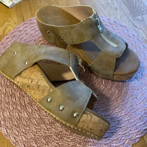 Beige sandaler med platå och dragkedja - Snygga beige tofflor med breda remmar i mocka-look, silverfärgade nitar och dragkedja framtill. Skorna har en hög platåsula i korkimitation och öppen tå, perfekt för en trendig sommarstil.