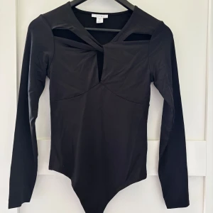 Svart långärmad body - Svart långärmad body med cut out-detalj vid halsen och twistad front. Sitter tight och har en bodyformad passform som ger en snygg siluett. 