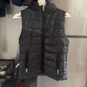 Svart dunväst från Mango  - Snygg svart dunväst från Mango med huva och två fickor med dragkedja. Västen är quiltad och har en rak passform. Perfekt att slänga över en hoodie för extra värme. Stängs med dragkedja framtill.