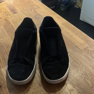 Svarta sneakers från Axel Arigato - Stilrena svarta sneakers från Axel Arigato med vit platt sula och diskret logga på sidan. Skorna har en minimalistisk design utan snörning och är tillverkade i mocka för en clean look. Perfekta för dig som gillar enkel och modern stil. De är använda ett par gånger men de har inga defekter.