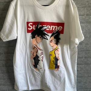 Vit Supreme t-shirt med anime-tryck - Vit t-shirt med Vegeta och Goku från Dragon ball Z. Klassisk rund hals och avslappnad passform. Perfekt för dig som gillar streetwear och anime. Helt ny oanvänd 