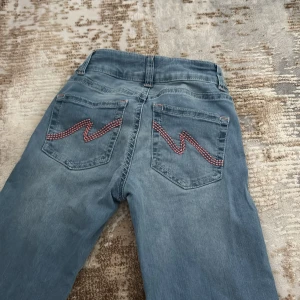Ljusblå bootcut jeans - ljusblå bootcut jeans från nelly med två knappar i midjan och coolt rödvitt broderi på bakfickorna. De är använda 3 gånger men har lite färg fläck på sig. Storlek 32
