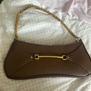 Brun axelväska med guldkedja från H&M - Snygg brun axelväska från H&M i skinnimitation med guldfärgade detaljer och kedja som axelrem. Väskan har en unik form, dragkedja upptill och ett stilrent metallspänne framtill. Perfekt för dig som vill ha en trendig och stilren accessoar.