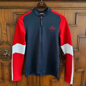 J.Lindeberg sporttröja med dragkedja - Sportig långärmad tröja från J.Lindeberg i marinblått, rött och vitt. Tröjan har hel dragkedja framtill, hög krage och logga på bröstet samt J.Lindeberg-tryck på ärmen. Tillverkad i polyester, perfekt för aktiva dagar och snygg streetstyle.