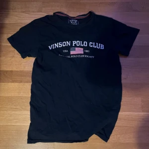 Mörkblå t-shirt Vinson Polo Club S - Mörkblå t-shirt från Vinson Polo Club med tryck framtill och amerikansk flagga. Klassisk rund halsringning och korta ärmar. Skön passform och mjukt bomullsmaterial, perfekt till jeans eller shorts.