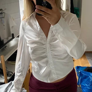 Vit croppad skjorta med knappar - Snygg vit skjorta med croppad passform och långa ärmar. Skjortan har knappar framtill och cool rynkdetalj längs bröstet som ger en unik look. Perfekt för dig som vill ha en stilren men ändå trendig vibe.