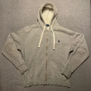Grå hoodie från Polo Ralph Lauren - Snygg ljusgrå hoodie från Polo Ralph Lauren med dragkedja och vit snörning i huvan. Klassisk broderad logga på bröstet                       Priset går o diskutera 