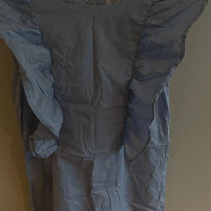 Ljusblå volangklänning från Lindex - Supersöt ljusblå klänning från Lindex med stora volanger längs axlarna och öppen rygg med knytband. Klänningen har resår i midjan för en bekväm passform och är tillverkad i mjuk bomull. Perfekt för dig som gillar en lekfull och trendig stil.