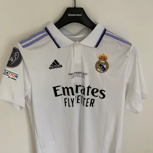 Real Madrid Champions League 2022 tröja - Real Madrid Champions League 2022 matchtröja i vitt från Adidas. Klassisk pikékrage med lila detaljer, klubbmärke och Adidas-logga på bröstet. Tryck med 'Emirates Fly Better' och Champions 14 på ryggen. Köpt från unisport.