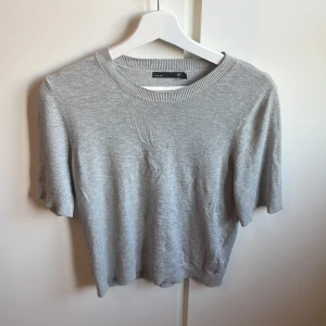 Grå stickad tröja från 157, S - Enkel och stilren grå stickad tröja från 157 med rund halsringning och korta ärmar. Tröjan har en mjuk känsla och ribbade detaljer vid hals, ärmslut och nederkant.