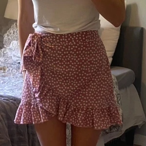 Rosa omlottkjol med volang och knyt - Supersöt kort omlottkjol i rosa med vita prickar. Kjolen har volangkant längs nederdelen och en knytning i sidan som ger en extra gullig touch. 