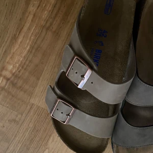 Beige sandaler från Birkenstock - Snygga beige sandaler från Birkenstock med två breda remmar och justerbara metallspännen i kopparfärg. Klassisk korksula och ovandel i mocka ger en skön och avslappnad vibe. Perfekta för varma dagar och chill outfits.