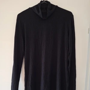 Svart ribbad polotröja i viskos - Svart ribbad polotröja i mjuk och stretchig viskosblandning. Tröjan har lång ärm och en hög krage som ger en clean och stilren look. Perfekt att styla med jeans eller kjol för en enkel men snygg outfit.