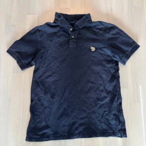 Pikétröja Paul Smith - Svart polo t-shirt från Paul Smith med klassisk krage och knappar framtill. På bröstet sitter en broderad zebra i färgglada ränder som ger en unik touch. T-shirten är gjord i bomull och har korta ärmar, perfekt för en stilren och avslappnad look.