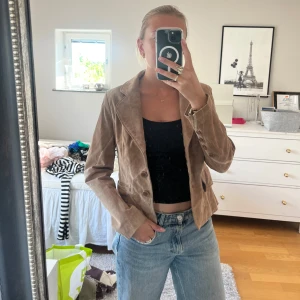 Brun mockakavaj med snygga detaljer - Säljer en brun kavaj i mocka med klassisk krage, knappar framtill och snygga sömdetaljer. Jackan har figursydd passform och veckad detalj bak som ger en cool siluett. Perfekt att styla med jeans för en cool och trendig look. Den är i nyskick och knappt använd! 