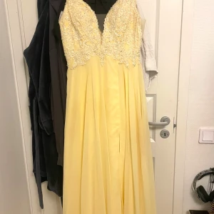 Gul långklänning med spets och pärlor - Säljer en elegant gul långklänning med v-ringad överdel dekorerad med spets och pärlor. Klänningen har smala axelband och ett flytande, plisserat kjolparti. Perfekt för bal eller festliga tillfällen. Gul klänning med paljetter och slits. Köpt från Beauty Queens i Södertälje. Använd 1 gång