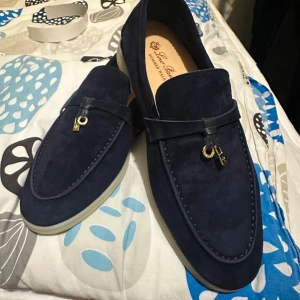 Blå mocka loafers från Loro Piana - Säljer ett par stilrena blå loafers från Loro Piana i mjuk mocka. Skorna har en dekorativ detalj framtill i form av en liten metallhängare och ljus sula. Perfekta för dig som gillar klassisk och elegant stil.