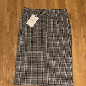 Rutig pennkjol från By Malene Birger - Säljer en klassisk rutig pennkjol från By Malene Birger i svart, vit och grå. Kjolen har hög midja och rak passform, perfekt till en stilren look. Dragkedja bak för enkel påklädning.