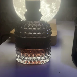 Valentino parfym  - Säljer en parfym i en unik, rund flaska med spetsmönstrad yta och svart lock. Flaskan har en gradient från klar till svagt rosa i botten och ett svart band runt mitten. Perfekt för dig som gillar snygg design på parfymflaskor.