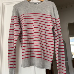 Grå- och röd randig tröja från J.Lindeberg - Säljer en stilren grå tröja från J.Lindeberg med röda horisontella ränder. Den perfekta tjocktröjan inför svensk sommar. Det står att den är i storlek L men skulle säga att den sitter som en M. 