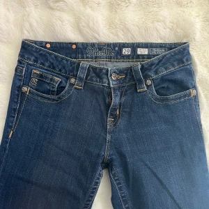 Mörkblå Miss Me jeans - Snygga mörkblå jeans från Miss Me med dekorativa knappar och detaljerade sömmar. Klassisk femficksmodell med logga på bakfickan och läderpatch i midjan. Perfekta för dig som vill ha ett par jeans med lite extra detaljer.