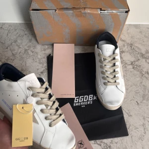 Vita sneakers från Golden Goose - Säljer ett par vita Golden Goose sneakers med beige snörning och grå stjärndetalj på sidan. Skorna har rund tå, platt sula och mörkblå häl. Klassisk och stilren design med märkets logga på sidan.