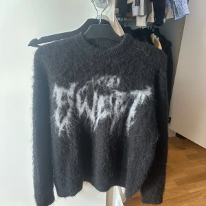 Svart stickad tröja från Sweet SKTBS - Cool svart stickad tröja från Sweet SKTBS med vit text framtill. Tröjan har lång ärm, rund halsringning och är gjord i ett fluffigt material som ger en unik look. Perfekt för dig som vill sticka ut med din stil.