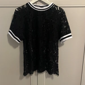 Svart spets t-shirt med vita ränder - Säljer en svart t-shirt i spets med vita ränder på ärmslut och krage. T-shirten är genombruten och har korta ärmar. Perfekt för dig som vill ha en cool och trendig look går att ha över ett linne under eller liknande toppen är i L men passar alla storlekar. 