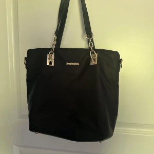 Svart axelväska från Juicy Couture - Säljer en svart axelväska med guldfärgade metalldetaljer och kedjor på handtagen. Väskan har en dragkedja på baksidan och är rymlig nog för vardagens alla prylar. Perfekt för dig som vill ha en stilren och praktisk accessoar.