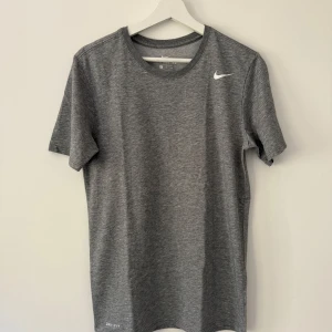 Grå Nike Dri-FIT t-shirt - Säljer en grå Nike t-shirt i Dri-FIT-material. T-shirten har rund halsringning, korta ärmar och en liten vit Nike-logga på bröstet. Perfekt för träning eller sportiga dagar.