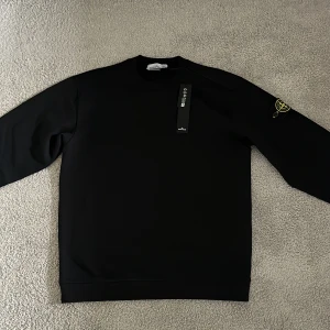 Svart sweatshirt från Stone Island - Säljer en svart sweatshirt från Stone Island med klassisk rund hals och ikonisk logotyp på vänster ärm. Tröjan har långa ärmar och en stilren, enkel design som passar perfekt till våren/sommaren. Passar runt S/M och är helt ny.