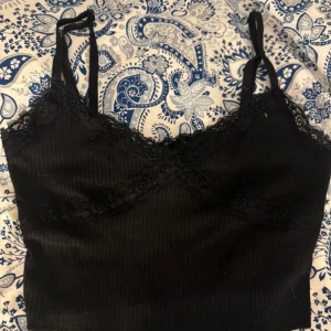 Svart croppad linnetopp med spets - Säljer en svart croppad linnetopp med tunna axelband och fina spetsdetaljer längs urringningen. Toppen är ribbad och har en tight passform, perfekt att styla till olika outfits.