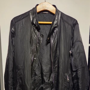 Svart jacka med skinndetaljer från Dolce & Gabbana - Säljer en svart jacka från Dolce & Gabbana med coola detaljer som dragkedjor och ståkrage. Jackan har långa ärmar och en snygg, stilren look med blandning av matta och blanka partier. Perfekt för dig som vill sticka ut.