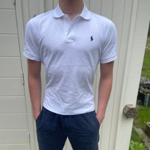 Vit pikétröja från Ralph Lauren - Säljer en klassisk vit pikétröja från Ralph Lauren med mörkblå broderad logga på bröstet. Tröjan har korta ärmar, krage och knappar framtill. Perfekt för en stilren och avslappnad look. Perfekt nu till sommaren. Den sitter riktigt bra, exakt som en piké ska sitta!! Den är s men passar men skulle säga att den passar mer som M. 